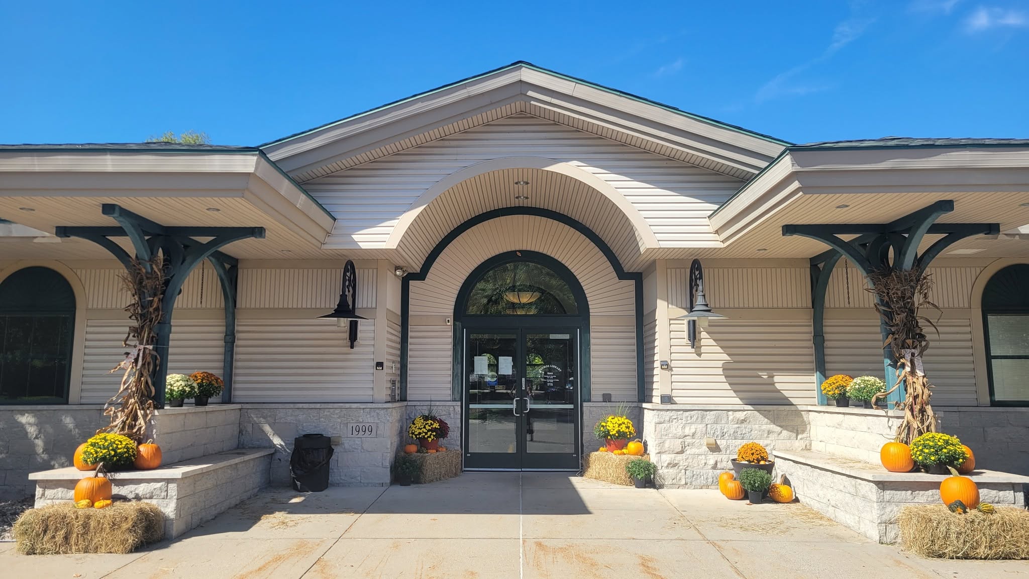 Image of the front of Patmos Library. The library's entrance is decorated with fall decor such as pumpkins, mums, corn stalks, and hay bales.